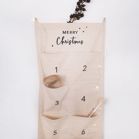 Adventskalender "Merry Christmas", 30 cm breit, von Eulenschnitt. Zum Befüllen, aus hellem Stoff mit zwei Schlaufen zum Aufhängen und aufgenähten Taschen. Abgebildet ist der Adventskalender mit Weihnachtsdeko hängend an einer weißen Wand.