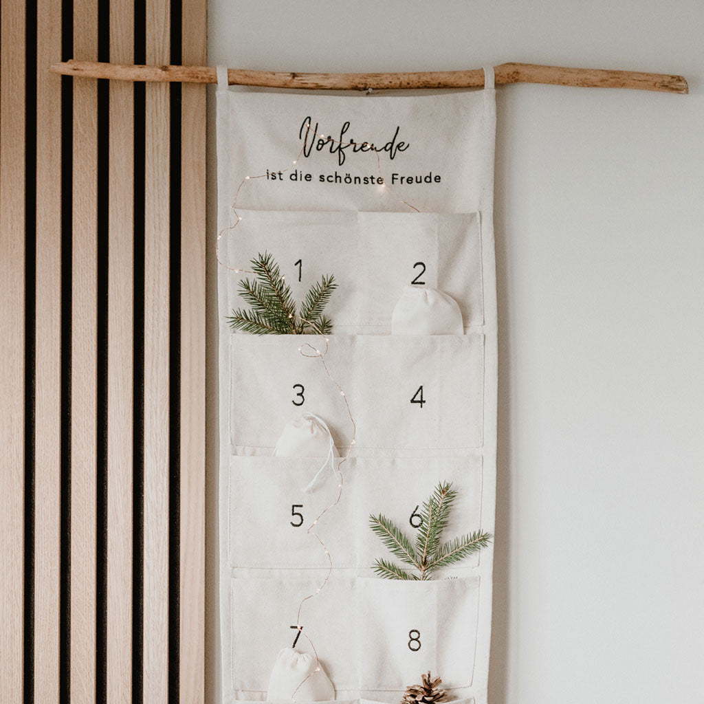 Adventskalender "Vorfreude ist die schönste Freude", 30 cm breit, von Eulenschnitt. Zum Befüllen, aus hellem Stoff mit zwei Schlaufen zum Aufhängen und aufgenähten Taschen. Abgebildet ist der Adventskalender hängend an einem Ast mit Weihnachtsdeko an einer weißen Wand mit Holzpanel.