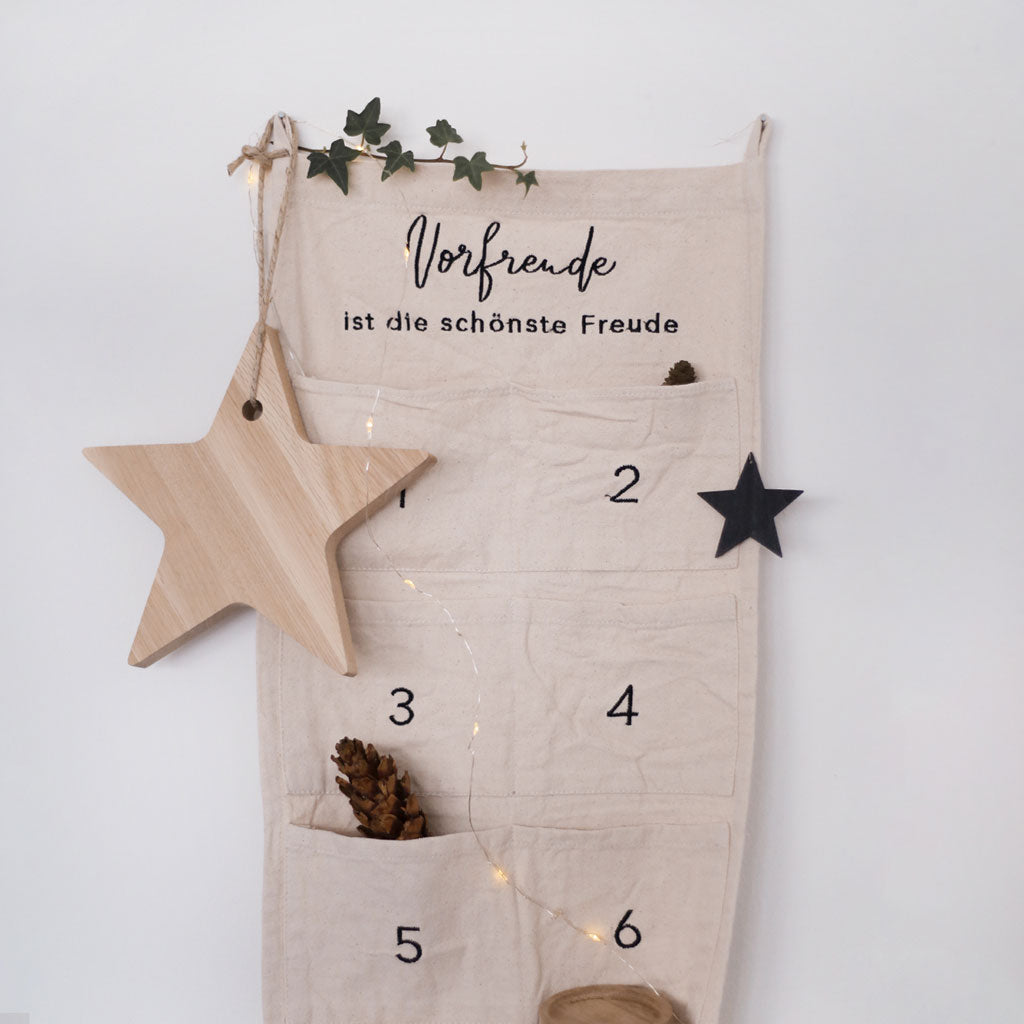 Adventskalender "Vorfreude ist die schönste Freude", 30 cm breit, von Eulenschnitt. Zum Befüllen, aus hellem Stoff mit zwei Schlaufen zum Aufhängen und aufgenähten Taschen. Abgebildet ist der Adventskalender hängend mit Weihnachtsdeko an einer weißen Wand.