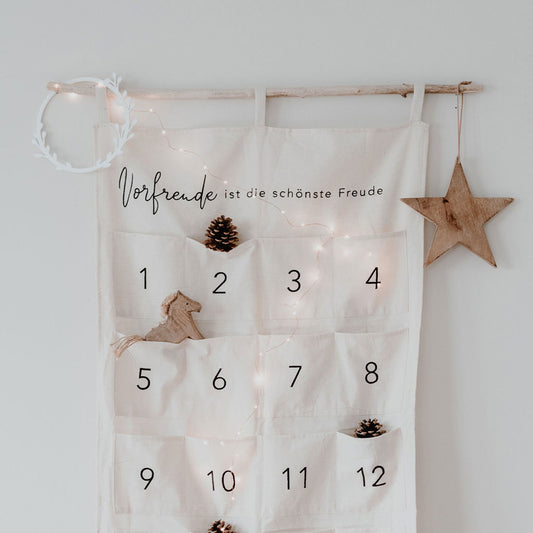 Adventskalender "Vorfreude ist die schönste Freude" von Eulenschnitt .Zum Befüllen, aus hellem Stoff mit drei Schlaufen zum Aufhängen und aufgenähten Taschen. Abgebildet ist der Adventskalender hängend an einem Ast mit Weihnachtsdeko an einer weißen Wand.